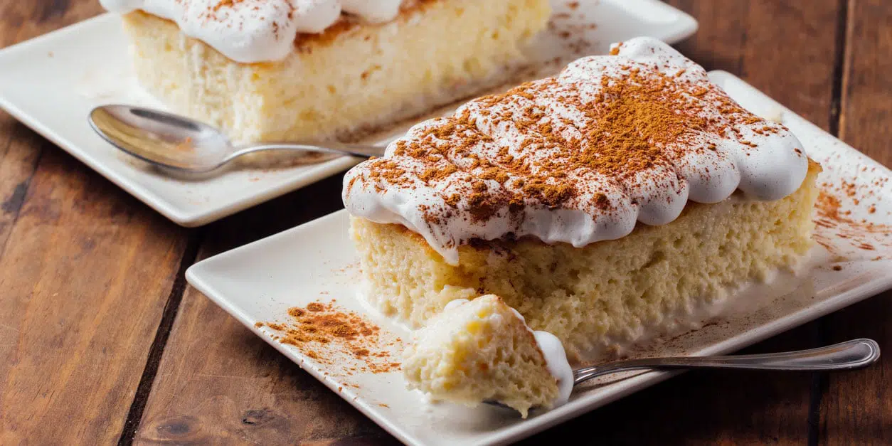 Tres Leches Cake Recipe for Imbolc 1 A fluffy slice of tres leches cake topped with whipped cream and sprinkled with a witchy blend of cinnamon, served on a square white plate with a dessert spoon on the side, ready to be