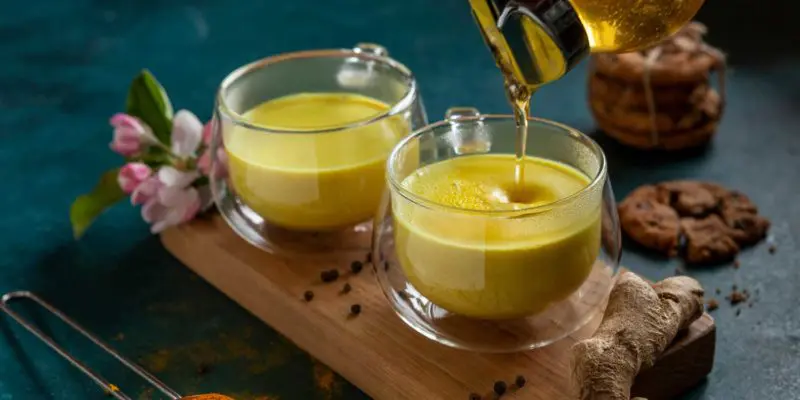 Golden turmeric tea being poured into a clear glass cup, with cookies and spices on a rustic kitchen countertop, creating a warm and healing atmosphere with a spiritual touch.