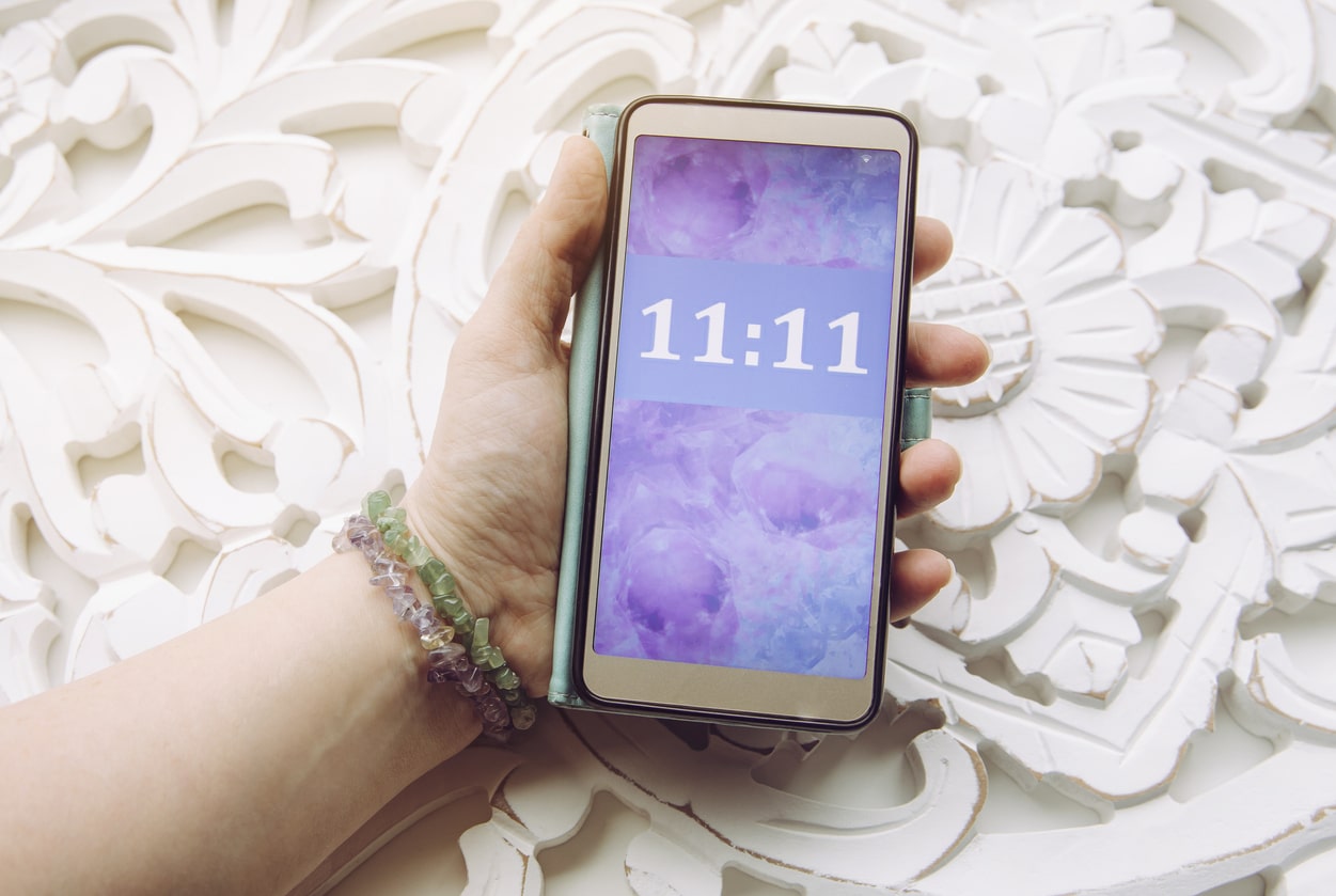 A person holding a smartphone showing 11:11 on a purple-themed screen, depicting an intricate white floral pattern with metaphysical symbols as the backdrop.