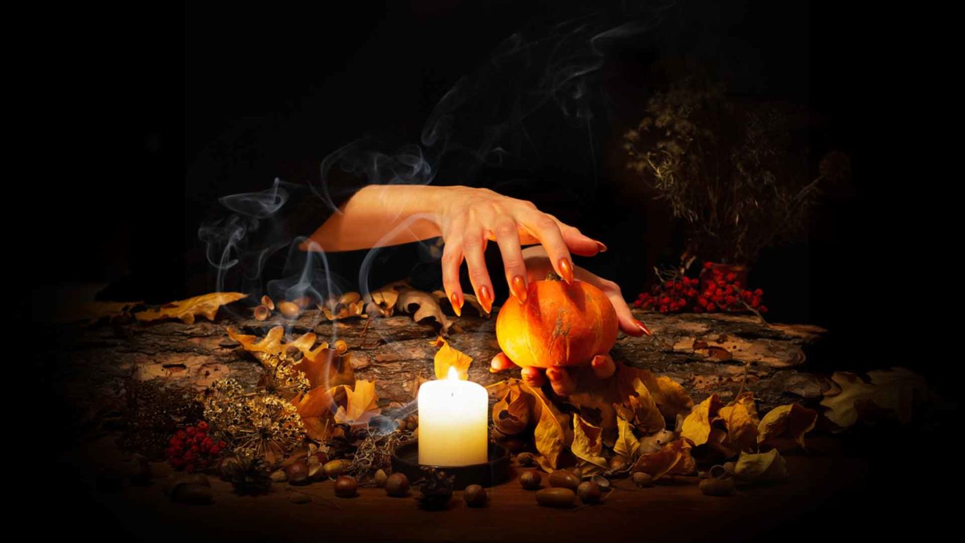 A mysterious hand appears over a small carved pumpkin surrounded by autumn leaves, acorns, and a glowing candle, creating an enchanting and witchy Halloween atmosphere.