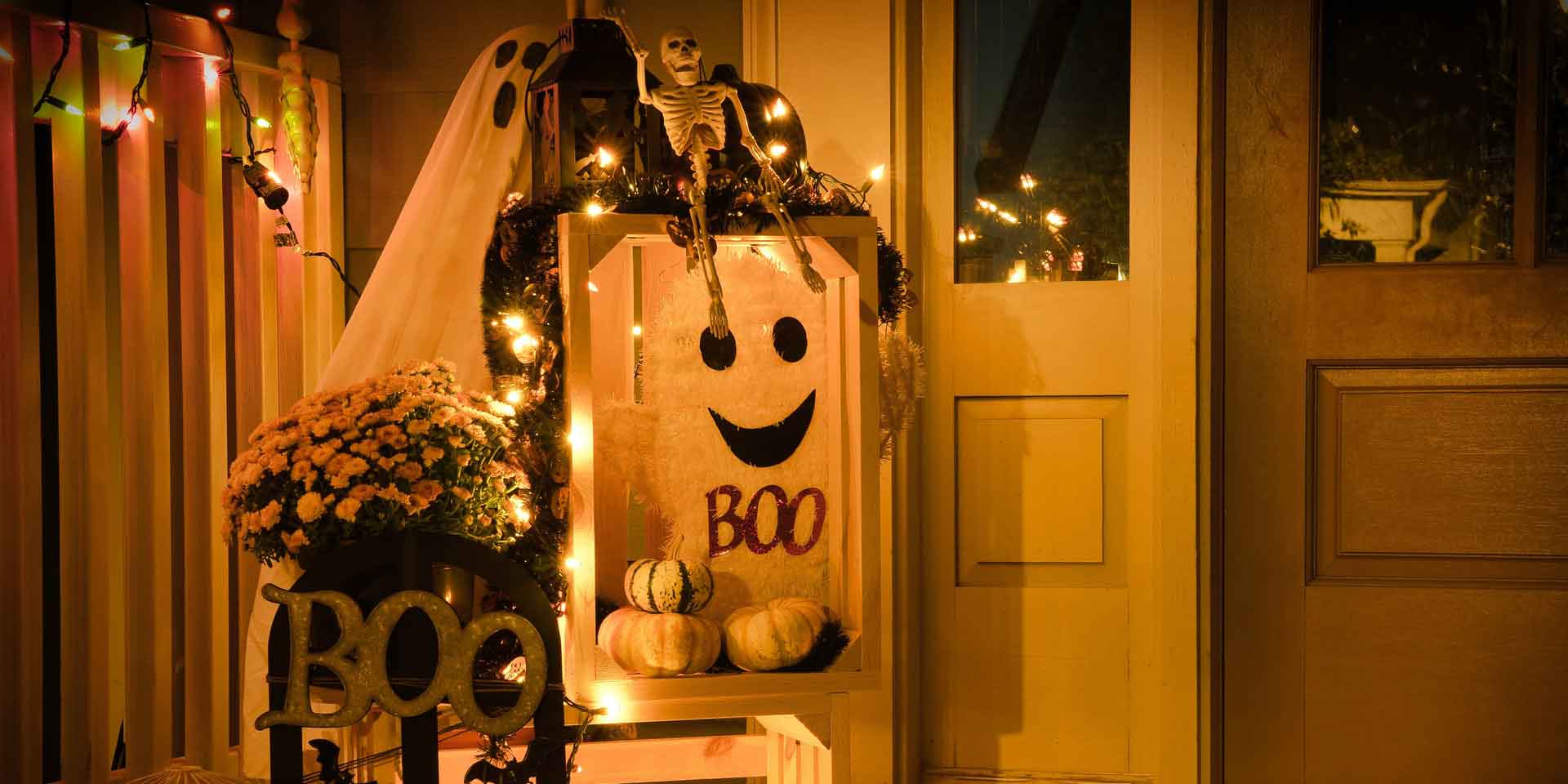 A warmly lit halloween-themed front porch decorated with a metaphysical ghost, a skeleton sitting atop a hay bale, twinkling lights, pumpkins, and a sign that says "boo".