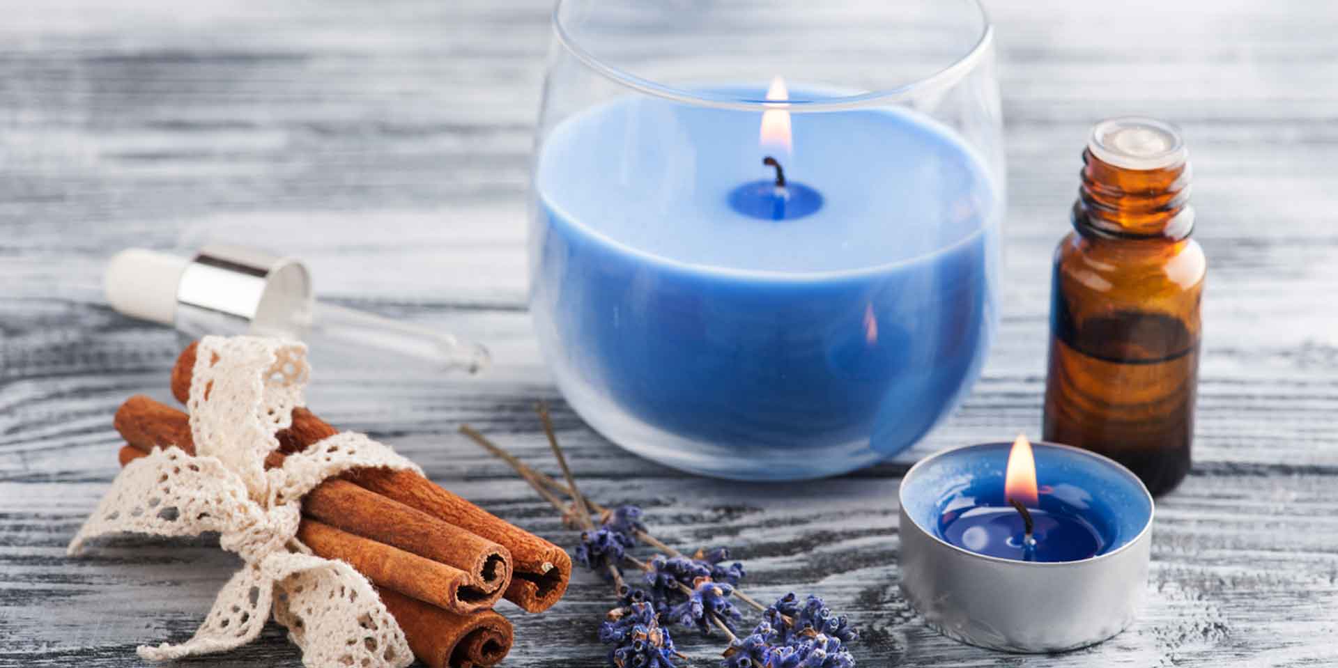 Two blue candles on a wooden table, surrounded by dried lavender, cinnamon sticks, and an essential oil bottle