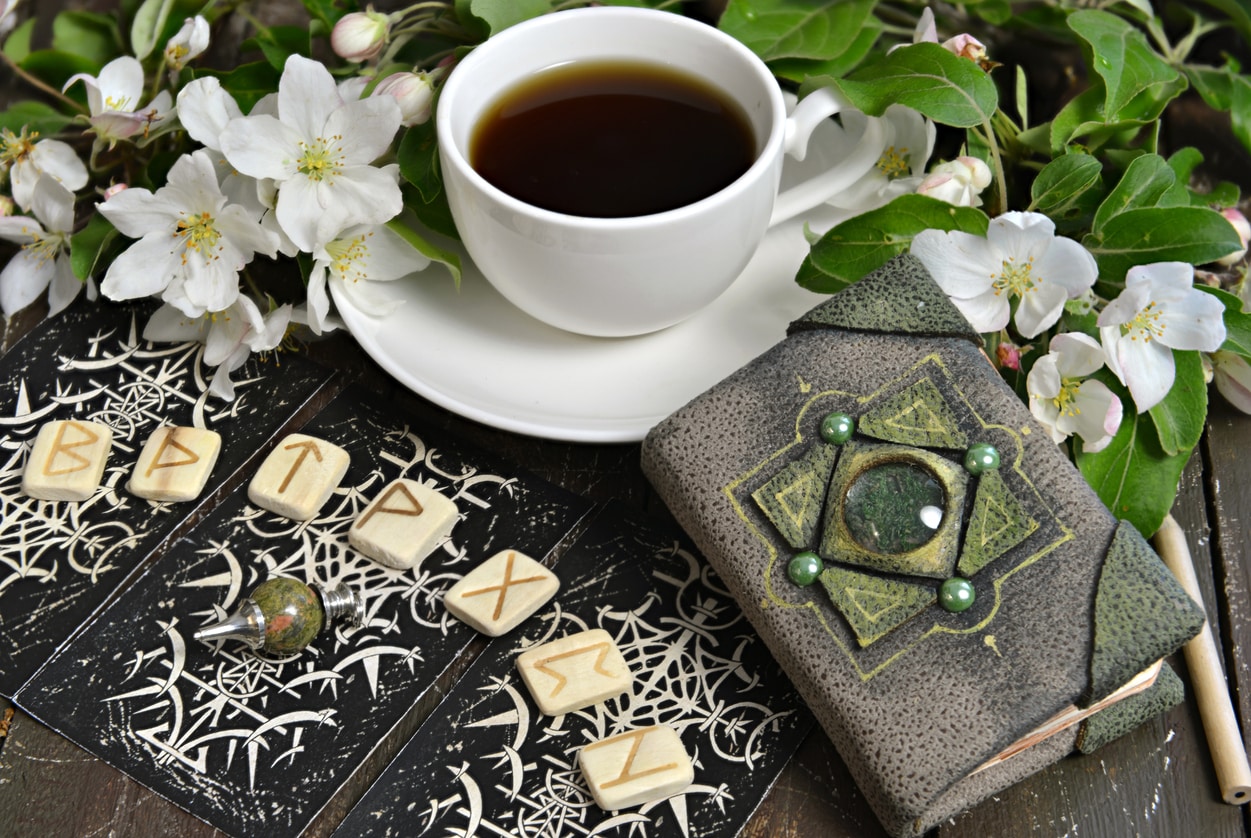 A cup of tea on a table with runes, flowers, and a leather journal