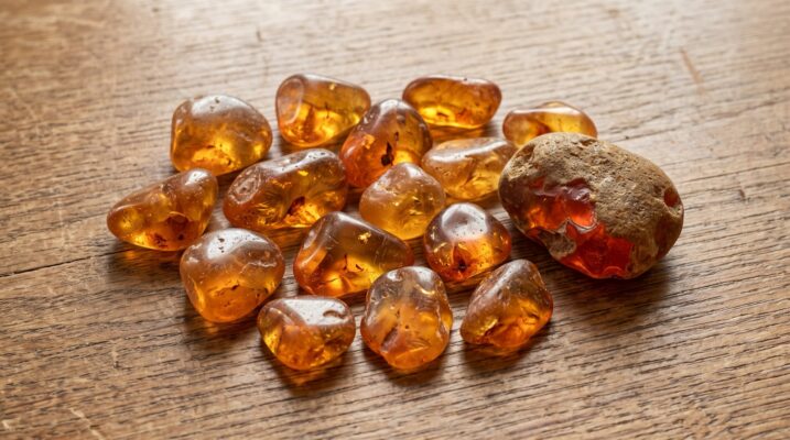 Amber tumbled stones and rough nugget Plentiful Earth | Global Spirituality Translucent honey and cognac tumbled amber pieces and one rough amber nugget on a wooden table