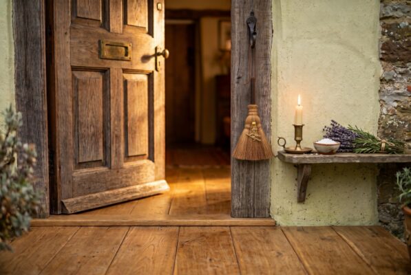 Besom hanging beside an open door Plentiful Earth | Global Spirituality A small besom broom of natural palm fiber with a gold witch charm hanging from an iron hook beside an open cottage door next to a shelf with a lit candle a bowl of salt and lavender and rosemary