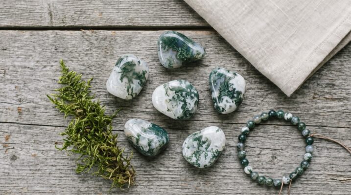Moss agate tumbled stones and bracelet flat lay Plentiful Earth | Global Spirituality Five tumbled moss agate stones with green dendritic inclusions beside a moss agate bead bracelet a sprig of moss and folded linen on weathered wood