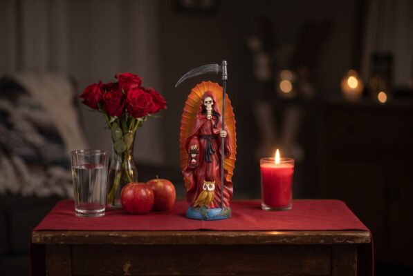 Santa Muerte statue on a home altar Plentiful Earth | Global Spirituality A Santa Muerte statue in a red robe with a scythe hourglass lantern and owl on a home altar with red roses apples a glass of water and a lit red candle