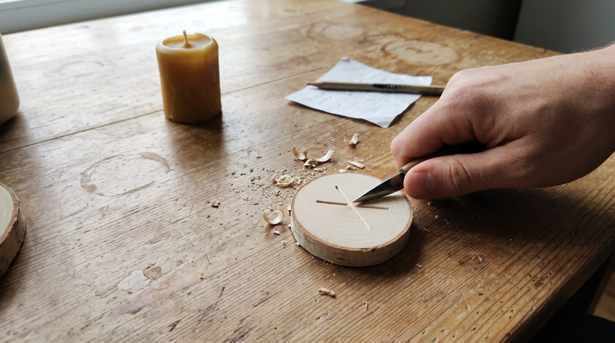 A hand carving a simple crossed rune into a round slice of birch wood with a small knife, with wood shavings and a beeswax candle on a wooden table.