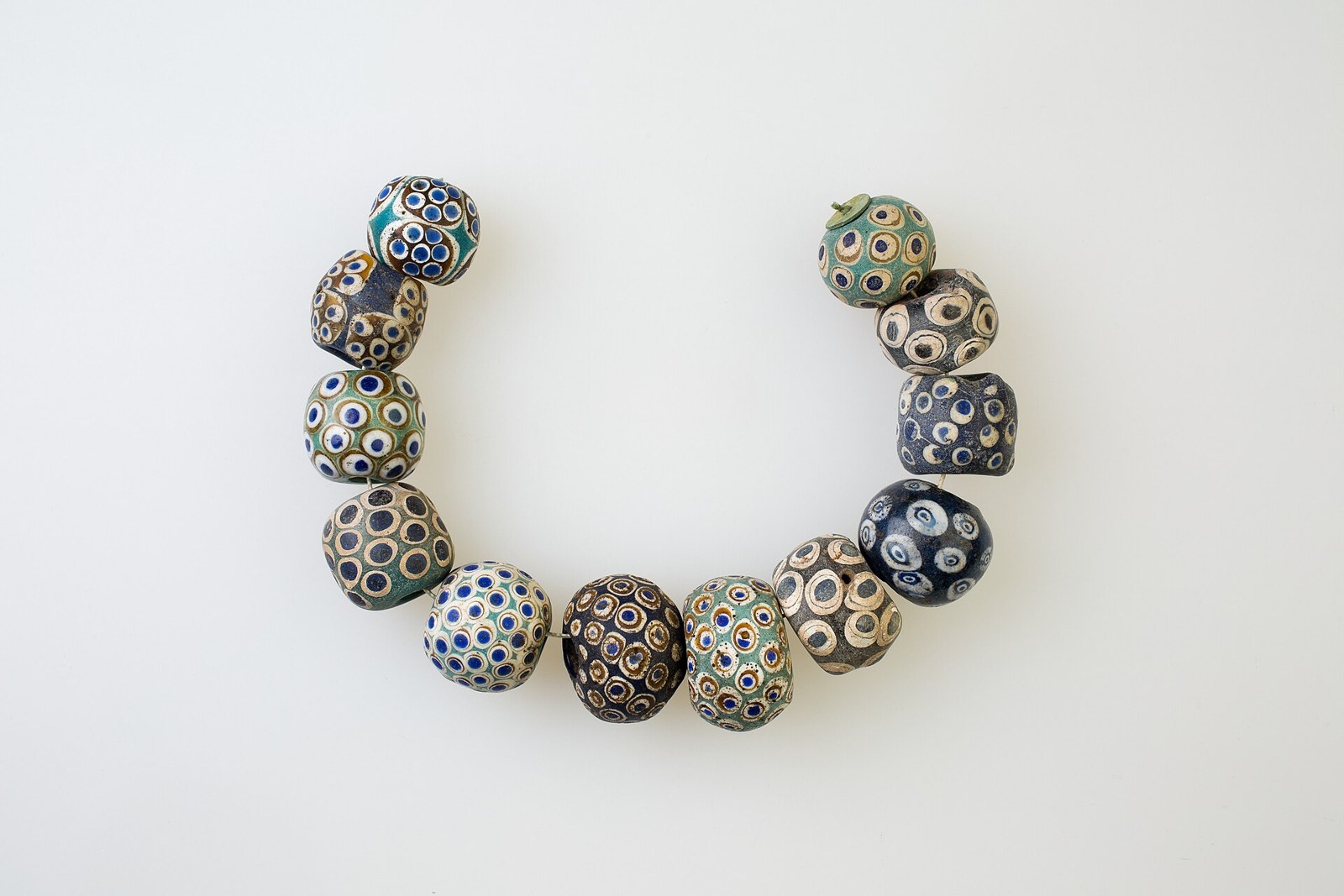 Egyptian glass eye beads Met Plentiful Earth | Global Spirituality Twelve ancient glass eye beads with dotted eye patterns arranged in a curve on a white background