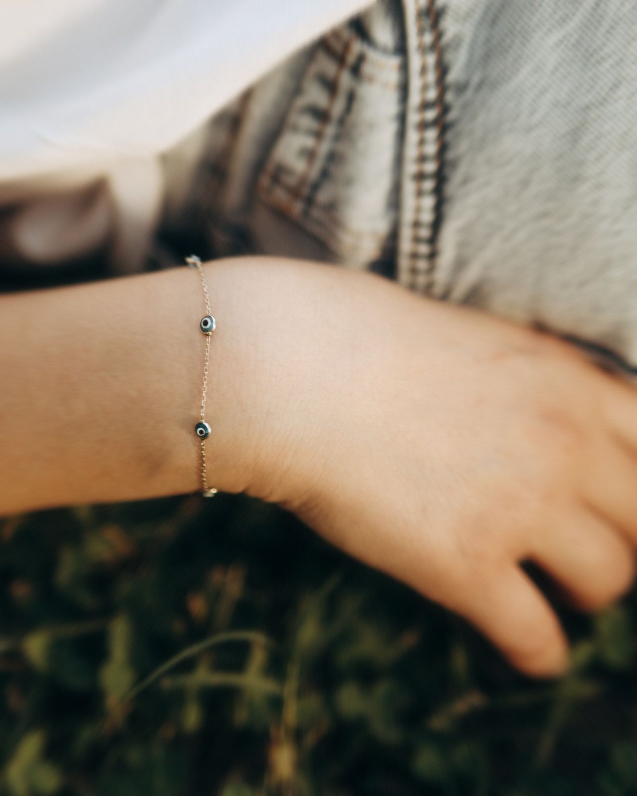 Evil eye bracelet on a wrist Plentiful Earth | Global Spirituality A delicate chain bracelet with small evil eye beads on a wrist, against a soft green outdoor background
