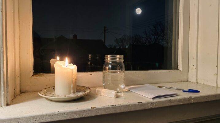 fullmoonritual Plentiful Earth | Global Spirituality A lit white pillar candle on a saucer a mason jar of water a selenite stick and folded notebook paper on a painted windowsill at night with a full moon in the window