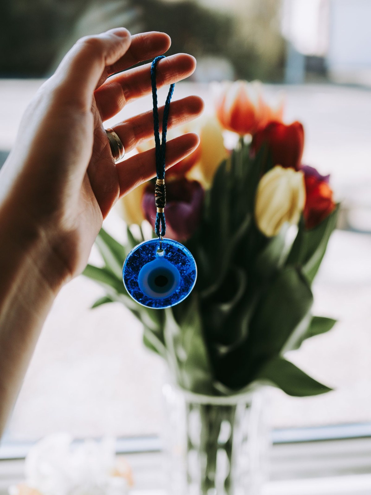 A hand holding up a single blue glass nazar bead on a cord in front of a vase of red and yellow tulips