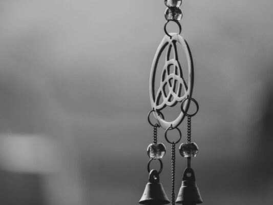 Triquetra wind chime Plentiful Earth | Global Spirituality Black-and-white close-up of a hanging metal wind chime shaped like a triquetra inside a circle, with small bells below