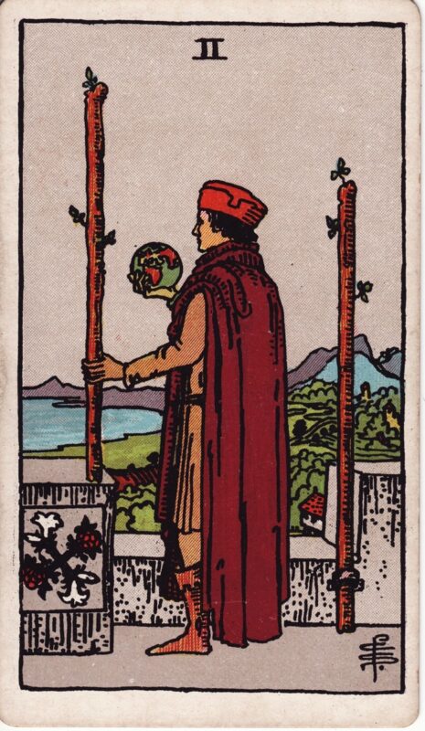 Two of Wands RiderWaiteSmith 1909 Plentiful Earth | Global Spirituality The Two of Wands tarot card from the 1909 Rider-Waite-Smith deck: a man in a red cap and cloak stands on a castle wall holding a small globe and a staff, looking out over the sea, with a second staff fixed in an iron ring beside him