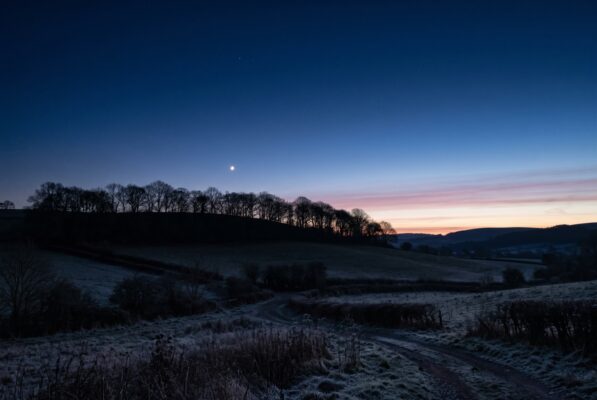 Moon visible in a deep blue twilight sky over a silhouette of bare trees along a snow dusted hillside